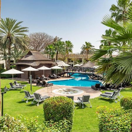 Le Meridien Dubai Hotel & Conference Centre Exterior photo The photo shows a serene outdoor scene featuring a swimming pool surrounded by lush greenery. There are several palm trees and shrubs providing a tropical atmosphere. Lounge chairs are arranged around the pool, along with shaded areas created by umbr