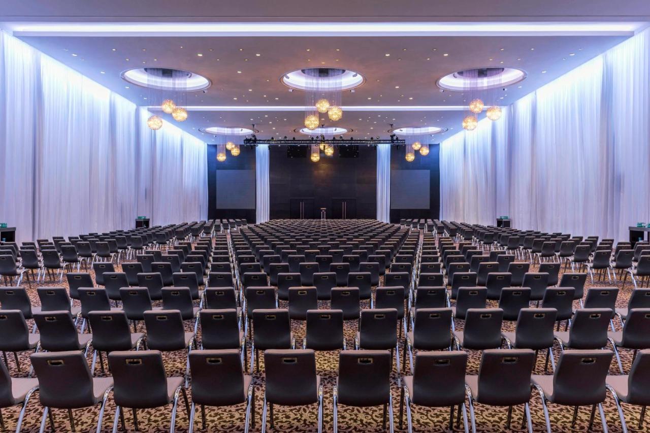Le Meridien Dubai Hotel & Conference Centre Exterior photo The photo shows a large conference or event hall set up with numerous rows of black chairs facing a staging area. The room features elegant lighting, with decorative pendants hanging from the ceiling. The walls are draped in white fabric, creating a 