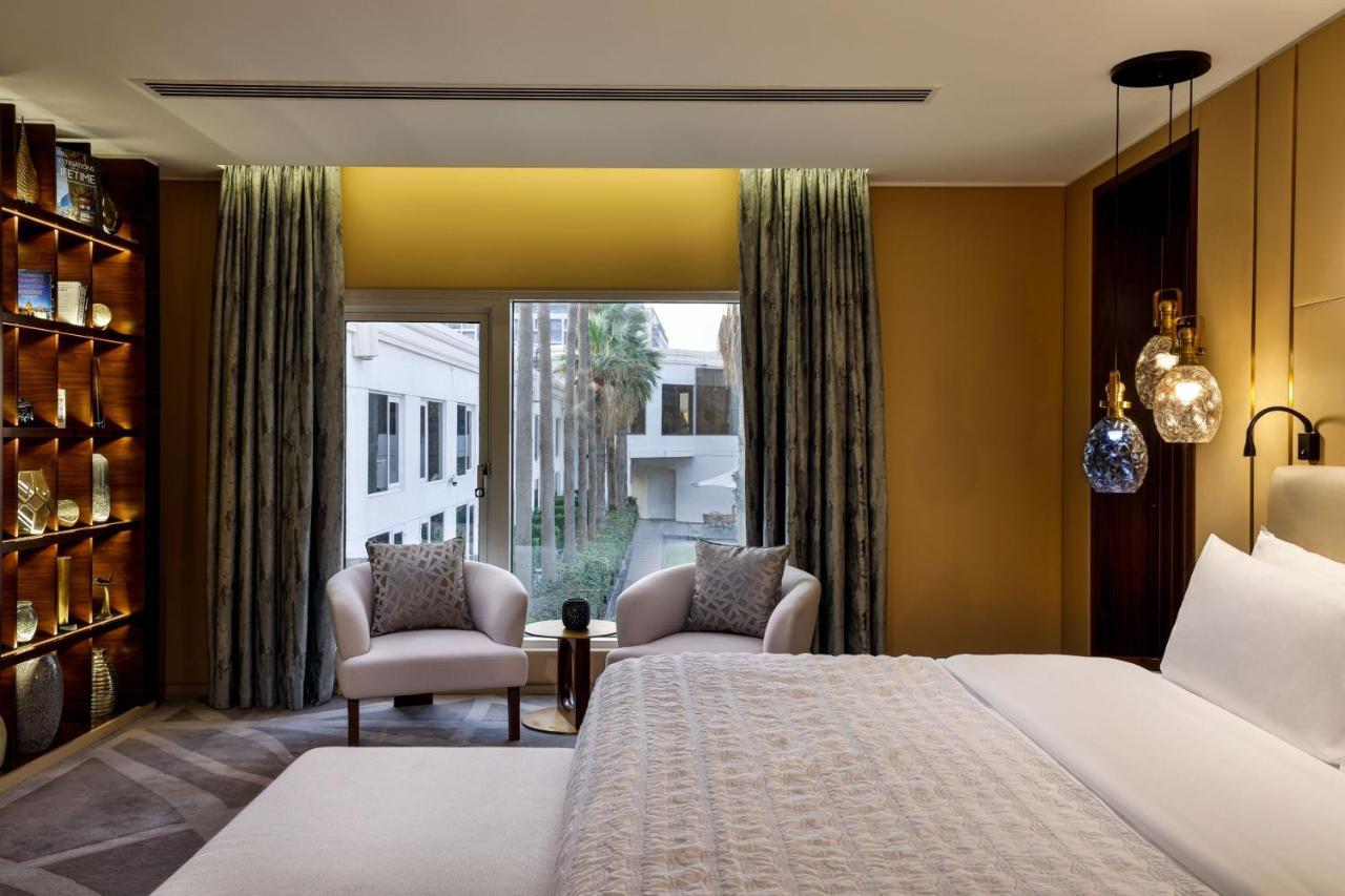 Le Meridien Dubai Hotel & Conference Centre Exterior photo The photo depicts a modern and elegantly designed hotel room. In the foreground, there is a large bed with a light-colored bedspread. On either side of the bed, there are stylish armchairs with decorative pillows. A small side table is positioned bet