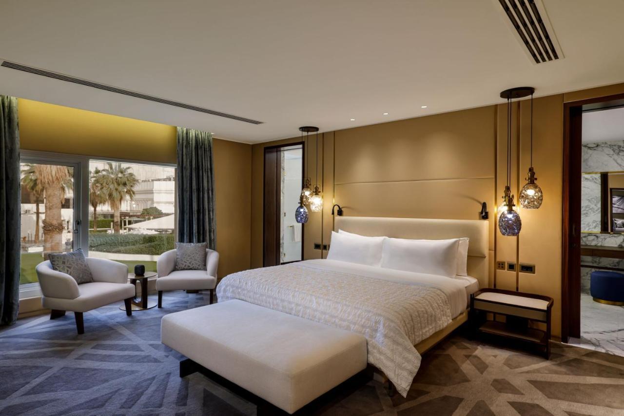 Le Meridien Dubai Hotel & Conference Centre Exterior photo The photo depicts a modern and elegantly designed hotel room. It features a large bed with soft white linens and a minimalist headboard. There are two stylish bedside lamps hanging on either side of the bed. In addition to the bed, there is a plush o