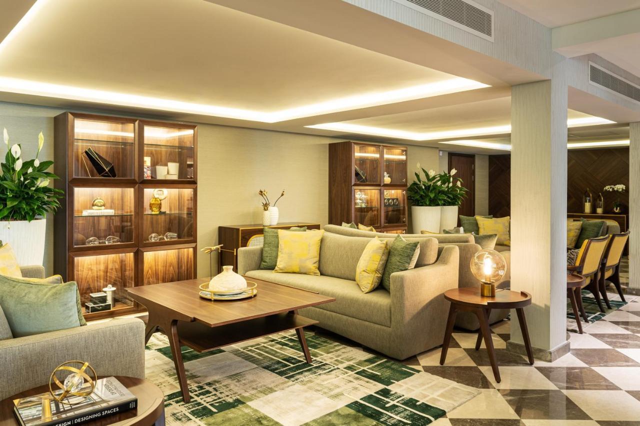 Le Meridien Dubai Hotel & Conference Centre Exterior photo The photo depicts a modern and elegantly designed lounge or living space. It features a light-colored sofa adorned with yellow and green cushions, and a coffee table in front of it. There are wooden shelves in the background, showcasing decorative it