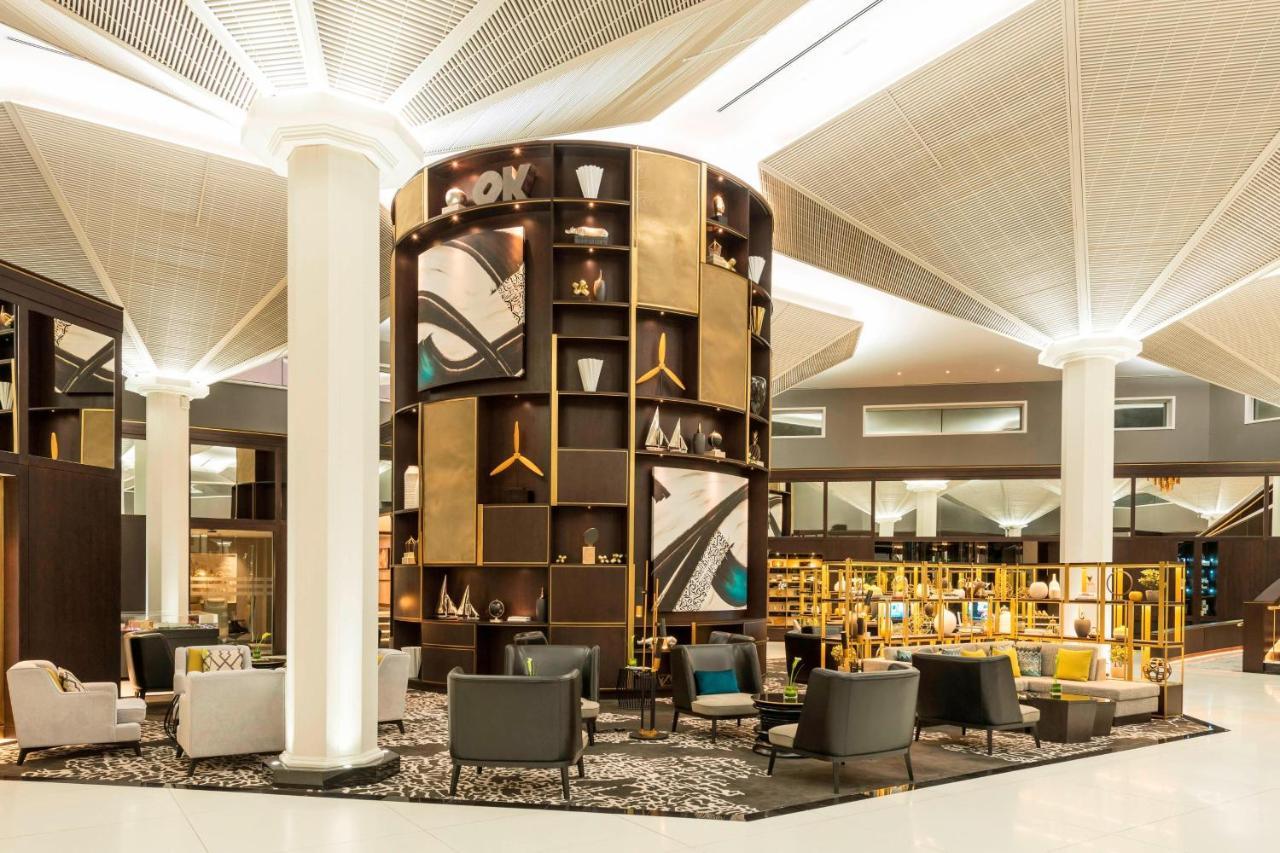 Le Meridien Dubai Hotel & Conference Centre Exterior photo The photo depicts a modern lounge or waiting area, characterized by a sleek and contemporary design. A prominent feature is a tall, circular shelving unit that displays various decorative items and art pieces, incorporating elements of gold and abstr