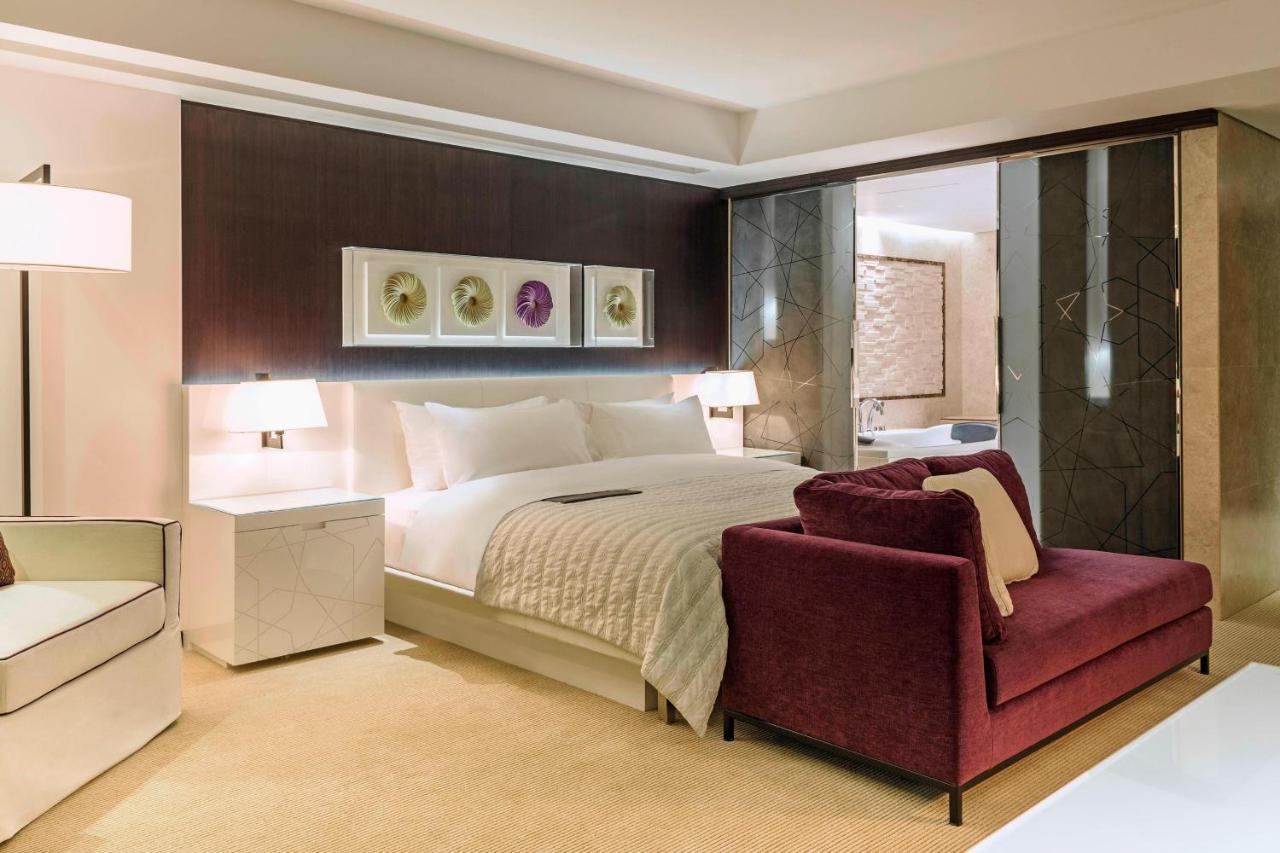 Le Meridien Dubai Hotel & Conference Centre Exterior photo The photo shows a modern hotel room featuring a spacious layout with a large bed, dressed in white linens, positioned against a dark accent wall. On either side of the bed are stylish bedside lamps. There is a burgundy armchair in front of the bed, p