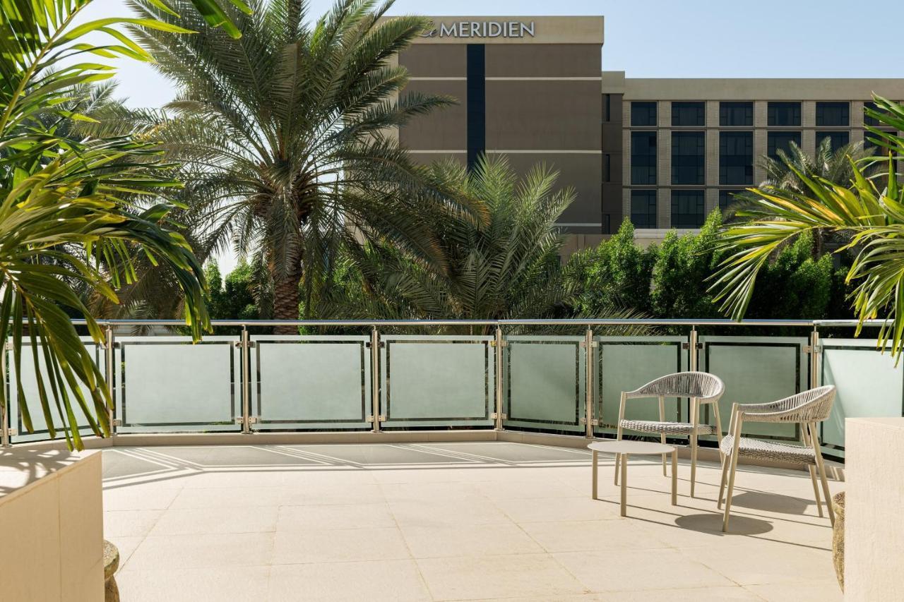 Le Meridien Dubai Hotel & Conference Centre Exterior photo The photo shows a balcony area with a paved floor and a railing. There are two white chairs positioned on the balcony. In the background, there is a building that appears to be part of a hotel, identifiable by the name "Le Meridien" on its exterior. 
