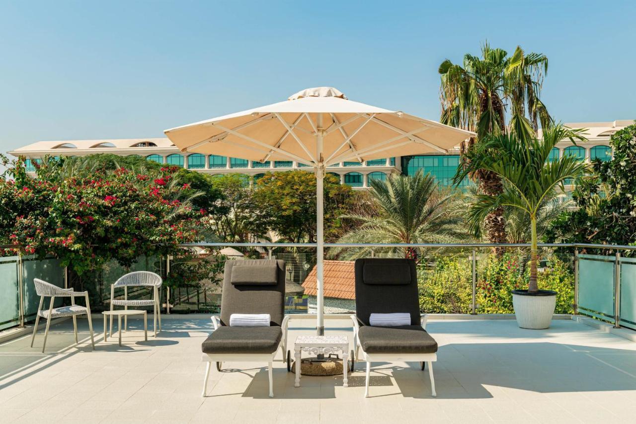 Le Meridien Dubai Hotel & Conference Centre Exterior photo The photo shows a relaxing outdoor space featuring two lounge chairs positioned beneath a large umbrella. The chairs are black and appear cozy, with white towels folded neatly on them. In the background, there are lush green plants, including palm tr