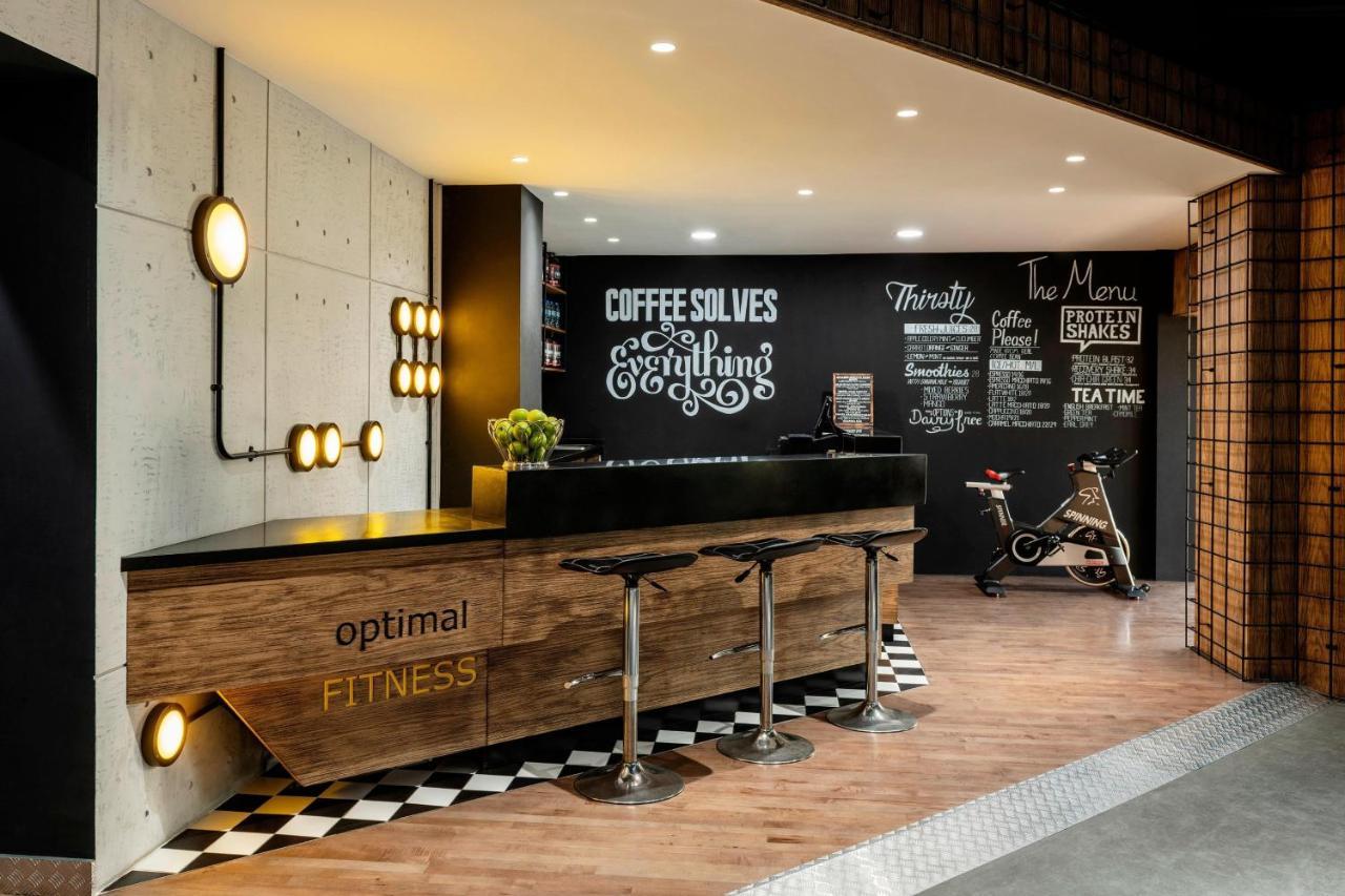Le Meridien Dubai Hotel & Conference Centre Exterior photo The photo showcases a modern fitness reception area. A sleek wooden reception desk with the words "optimal FITNESS" is positioned prominently. Behind the desk, a black wall features motivational phrases and chalkboard-style text, including "COFFEE SO