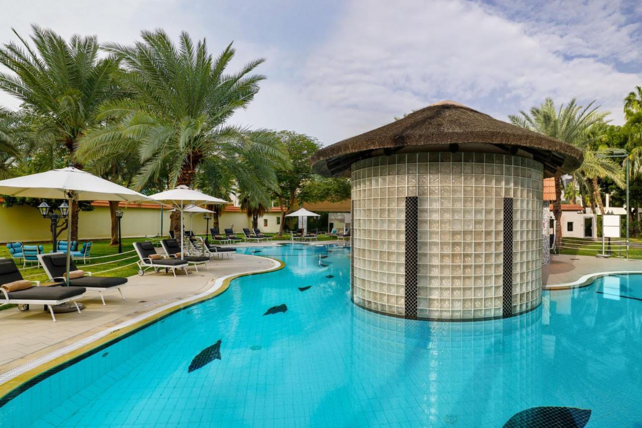 Le Meridien Dubai Hotel & Conference Centre Exterior photo The photo shows a luxurious pool area surrounded by palm trees. The pool has a unique design with a circular, glass-brick structure in the center, which may serve as a bar or lounge. There are lounge chairs placed around the pool, and several white u