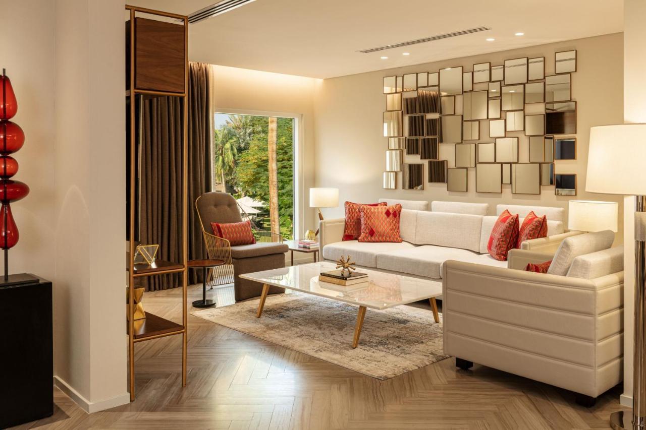 Le Meridien Dubai Hotel & Conference Centre Exterior photo The photo shows a modern living room interior. It features a white sofa adorned with red accent pillows, alongside a comfortable armchair in a muted color. A coffee table with a glass surface is positioned in front of the sofa, sitting on a light-col