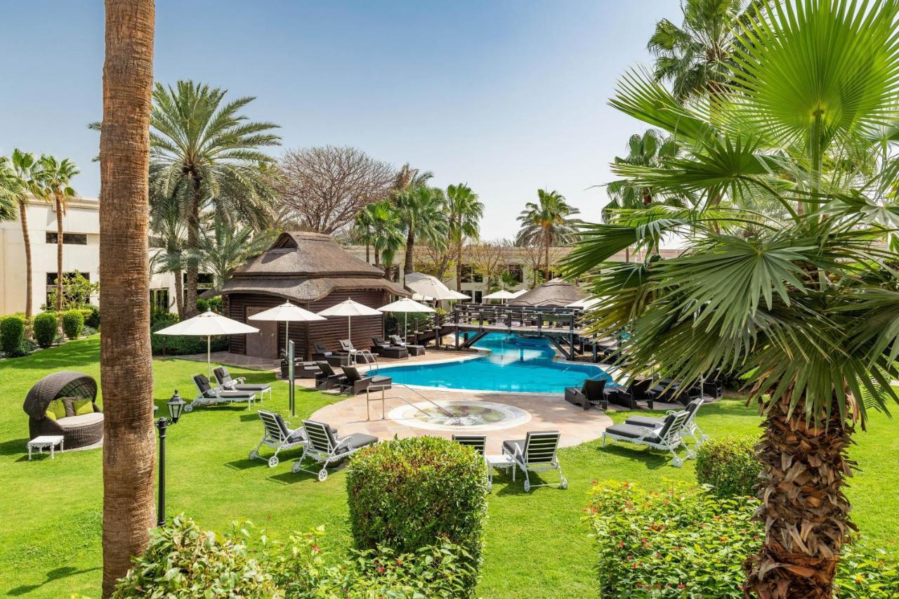 Le Meridien Dubai Hotel & Conference Centre Exterior photo The photo shows a serene outdoor scene featuring a swimming pool surrounded by lush greenery. There are several palm trees and shrubs providing a tropical atmosphere. Lounge chairs are arranged around the pool, along with shaded areas created by umbr