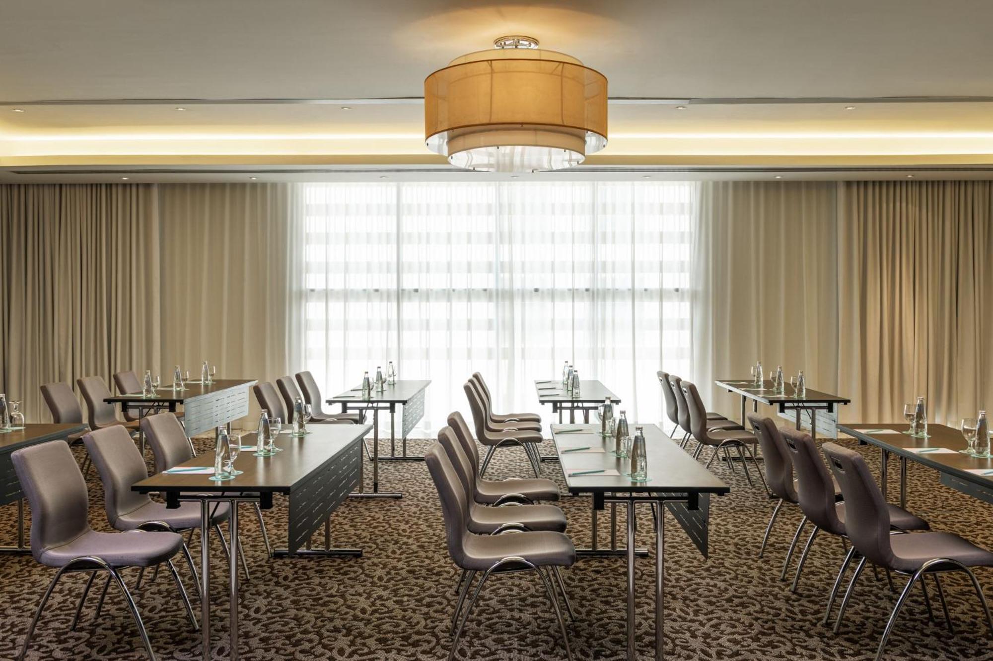 Le Meridien Dubai Hotel & Conference Centre Exterior photo The photo shows a conference room set up for meetings or presentations. There are several rows of tables with chairs arranged in a classroom style. Each table has a water bottle, suggesting that it is prepared for participants. The room is well-lit, 