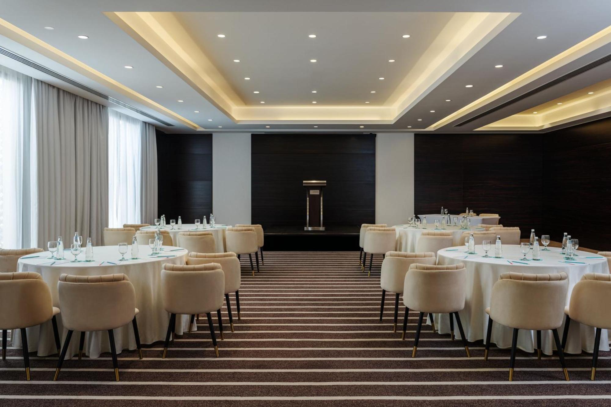 Le Meridien Dubai Hotel & Conference Centre Exterior photo The photo shows an elegantly designed conference or banquet room. There are round tables covered with white tablecloths, each accompanied by plush, light-colored chairs. The flooring features a striped pattern. The room is well-lit with recessed ceil