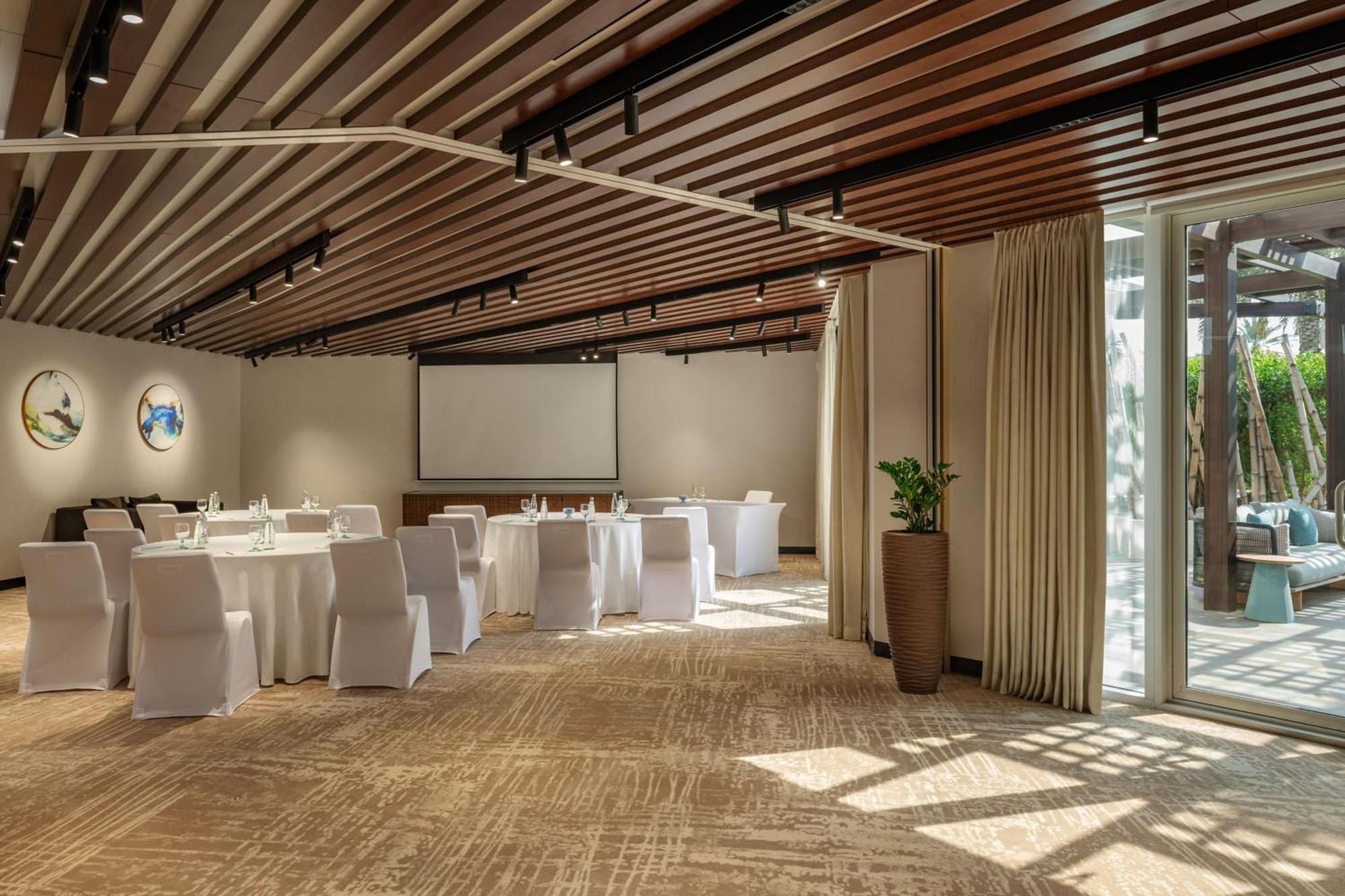 Le Meridien Dubai Hotel & Conference Centre Exterior photo The photo shows a modern, well-lit conference room or meeting space. The room features a high ceiling with wooden beams and has several tables set up in a semicircular arrangement, each covered with white tablecloths. There are white chairs around th