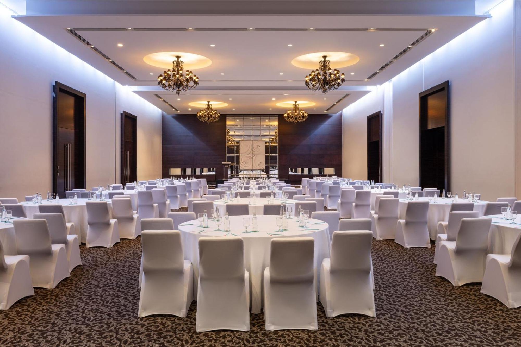 Le Meridien Dubai Hotel & Conference Centre Exterior photo The photo depicts an elegant banquet hall setup. The room features several round tables covered with white tablecloths, surrounded by white chair covers. The decor includes stylish chandeliers hanging from the ceiling, providing a warm and upscale am